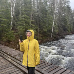 femme en parka jaune sur un pont. l'eau est forte en courant, la femme tient un micro